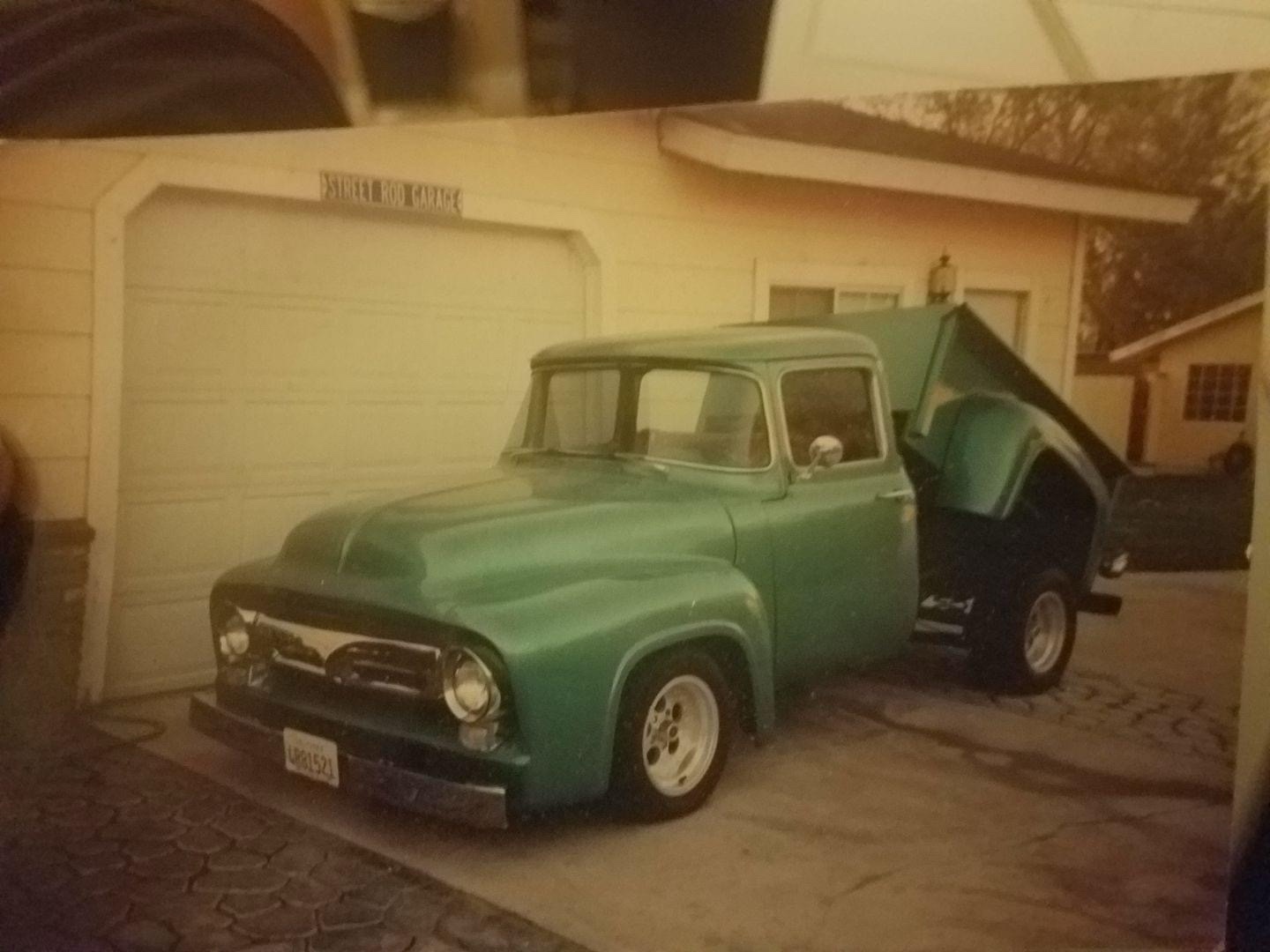 Green Ford F-100 with its bed raised, parked outside a garage.